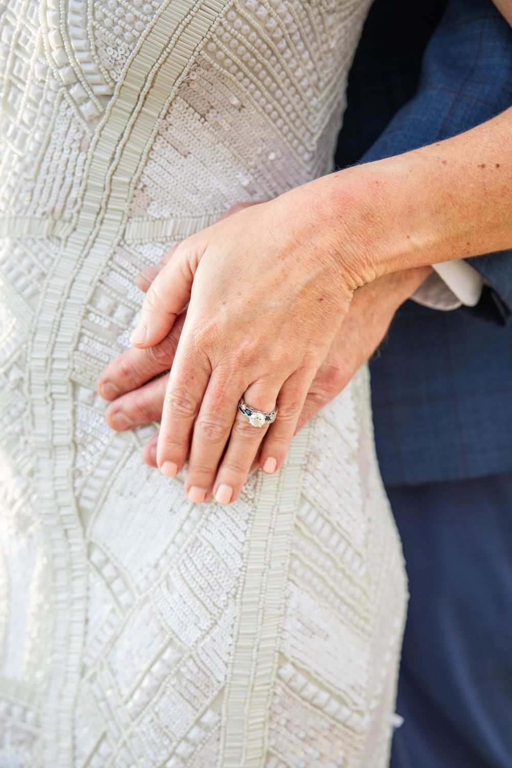 Close-up of hands wearing engagement ring resting on white lace dress fabric against navy background.
