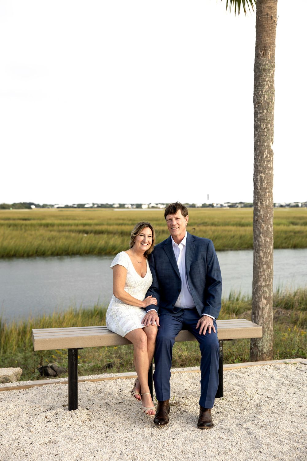 A couple sits together on a wooden bench overlooking a coastal marsh landscape on a sunny day.