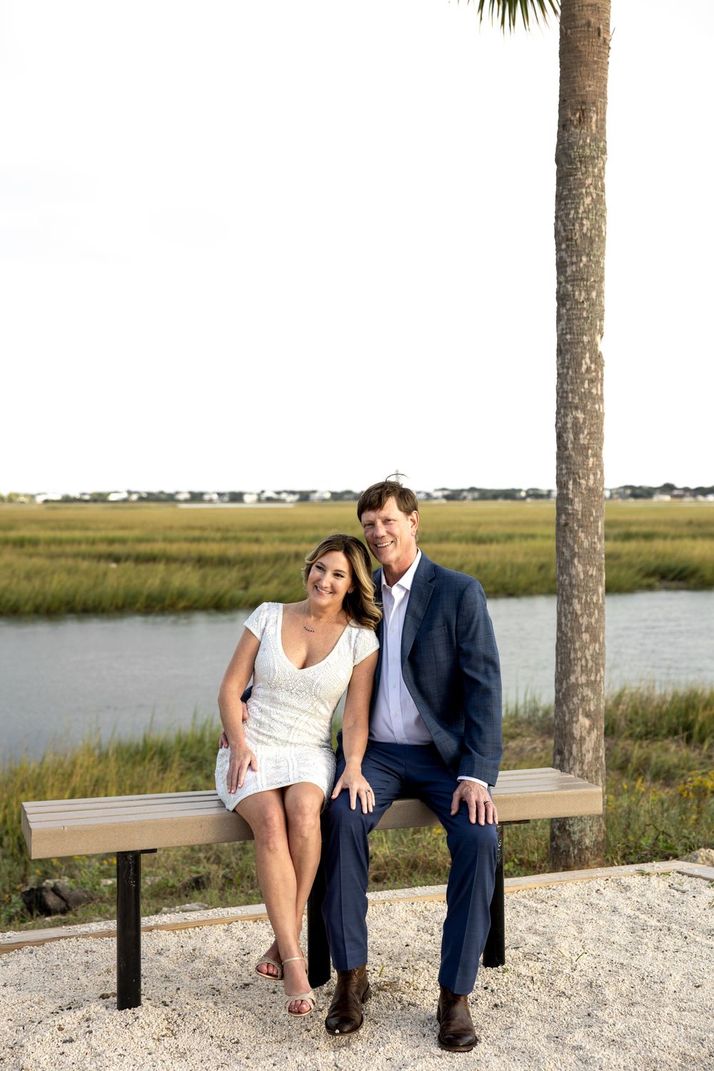 A couple sits together on a bench by the marsh at sunset with a palm tree overhead and water in the background.