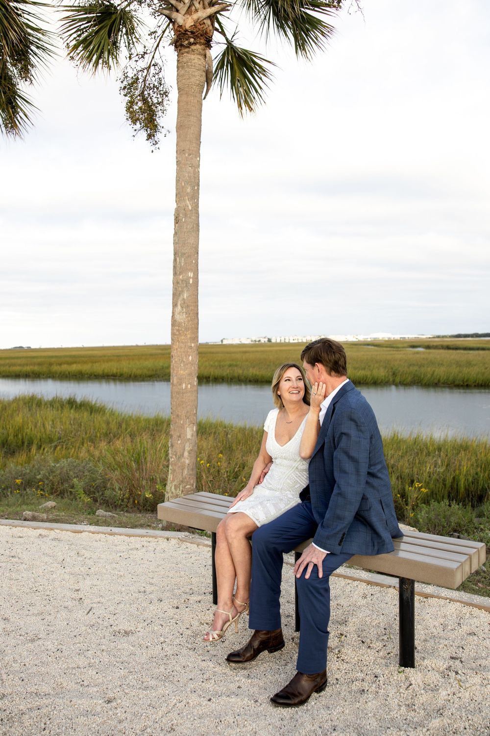 Couple sits together on wooden bench along coastal marsh with palm trees and water views in background.