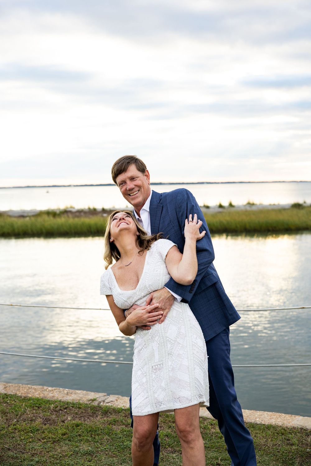 A joyful couple shares a romantic moment by the waterfront during sunset, with marsh grass and water in the background.