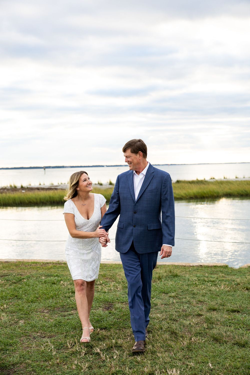 A couple in wedding attire shares a playful moment on grassy waterfront during their outdoor photo session.