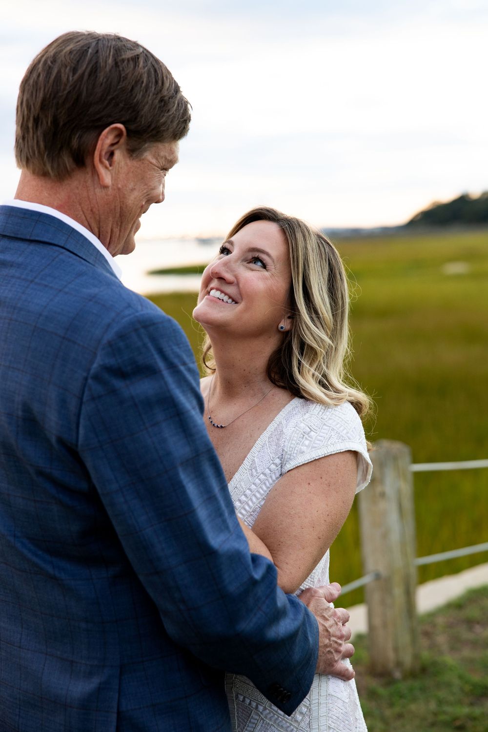 A couple shares a romantic moment together in a rustic outdoor setting with wooden fencing and green fields.
