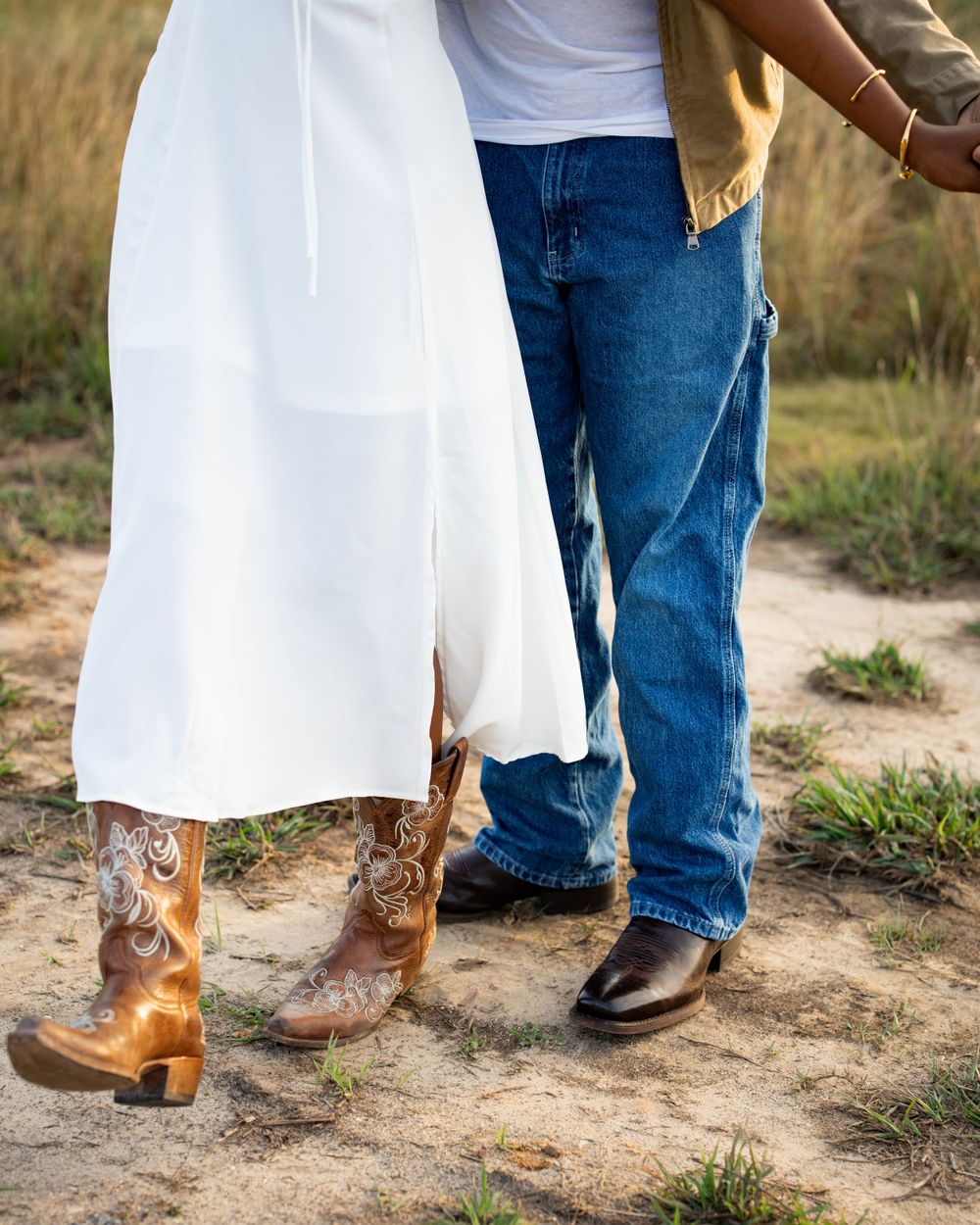 A pair of decorative brown cowboy boots next to casual black boots on a dirt path, with white dress fabric between them.