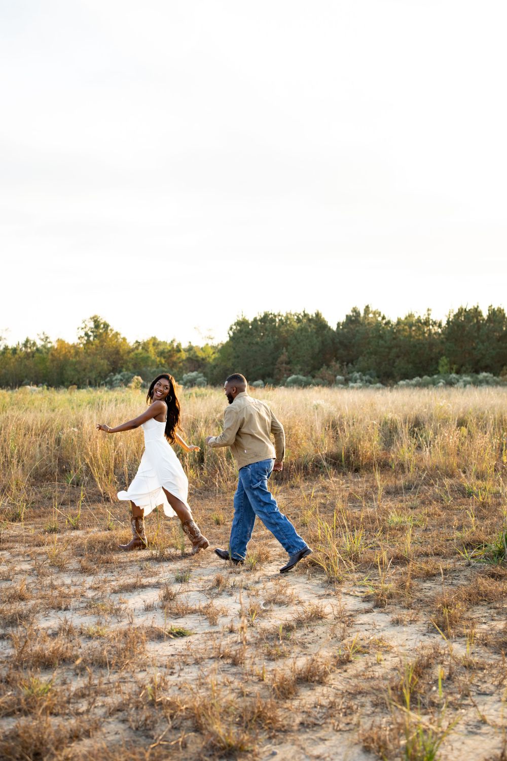 A couple dances together in a golden field during sunset, with her white dress swaying as they move.