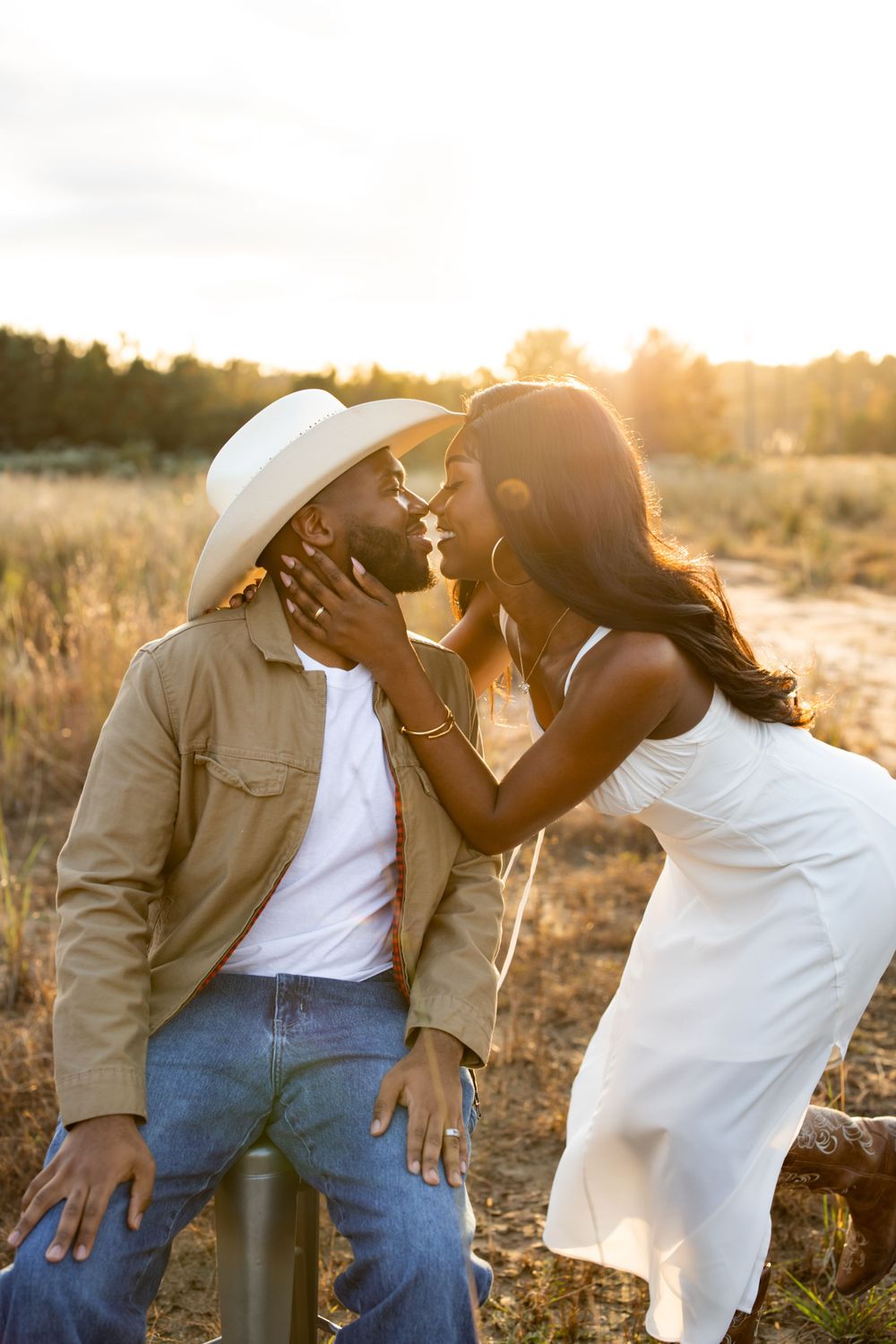 A romantic couple shares an intimate moment in a field at golden hour, with one wearing a white hat and tan jacket.