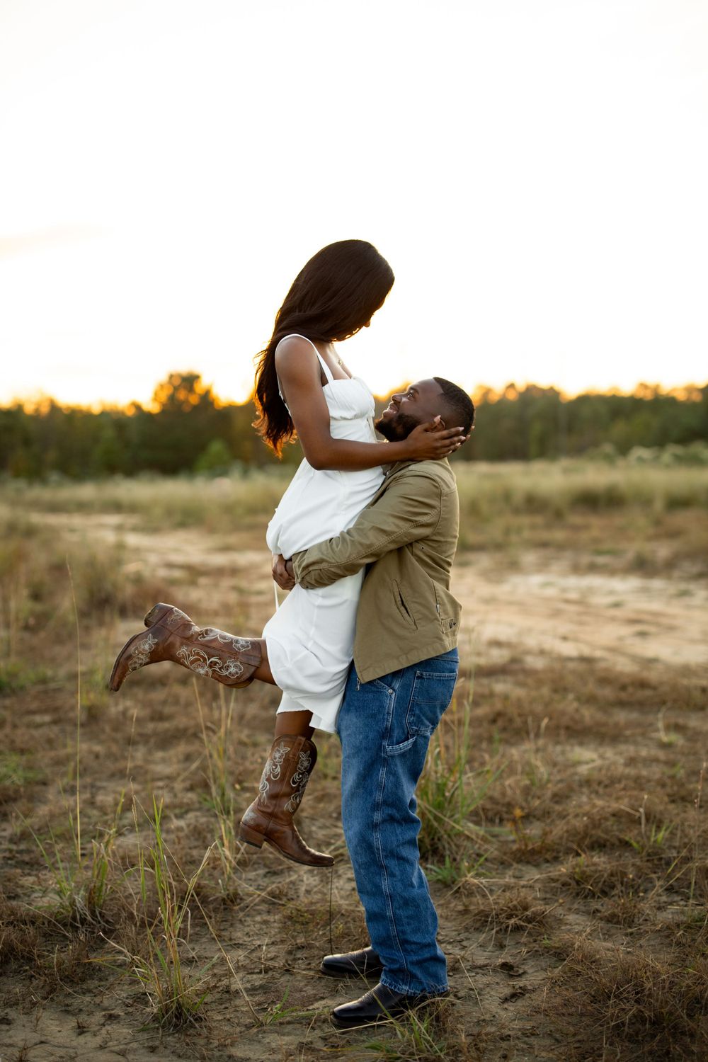 A couple embraces in a romantic sunset moment outdoors, with the woman wearing a white dress and boots in a rural setting.