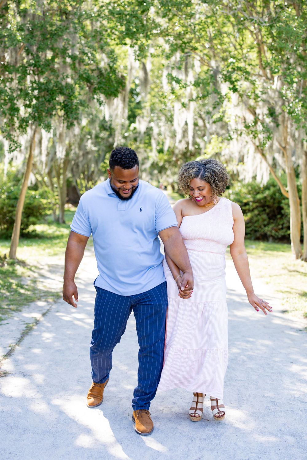 A couple walks hand in hand down a scenic path with Spanish moss hanging overhead.