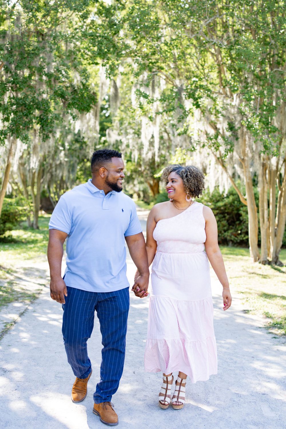 A couple takes a romantic stroll down a sunlit path surrounded by trees.