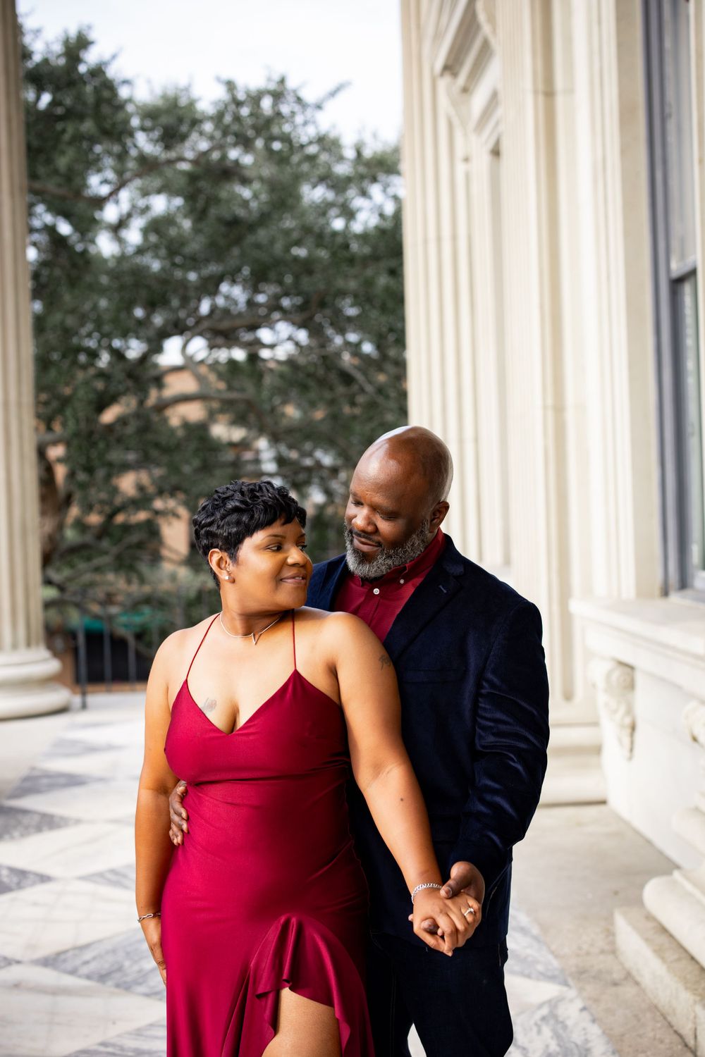 Couple in burgundy dress and navy blazer sharing romantic moment by classical building.