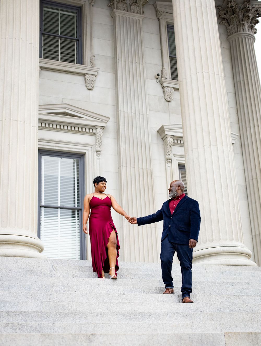 Two people in formal attire holding hands on white columned building steps.