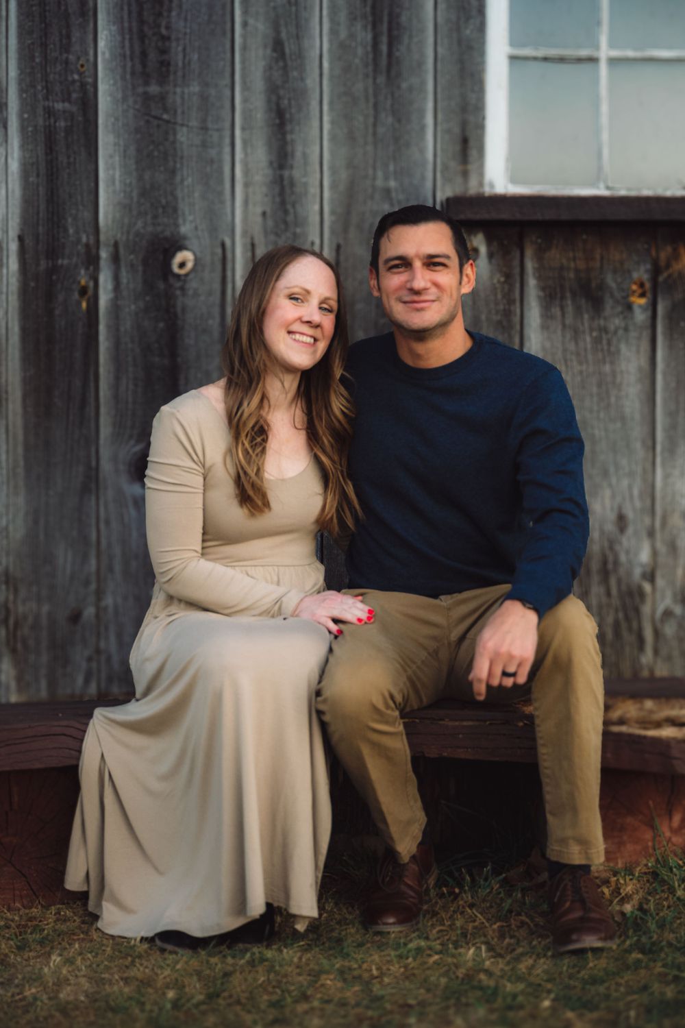 Couple sitting on wooden bench against rustic barn wall.