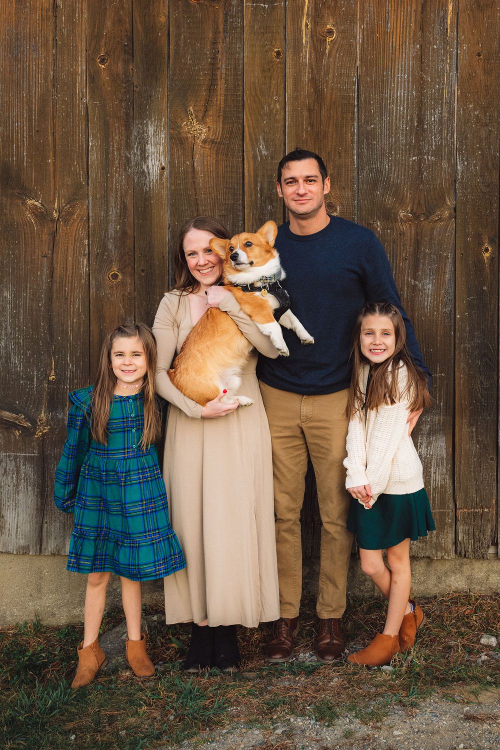 Family poses against rustic wooden fence with their corgi dog on an autumn day.