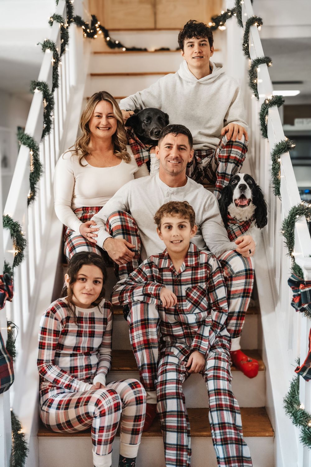 Family in matching plaid Christmas pajamas pose on white staircase with garland.