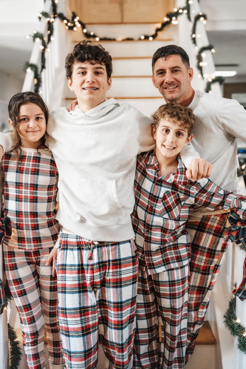 Family wearing matching plaid Christmas pajamas poses together on stairs decorated with holiday garland.