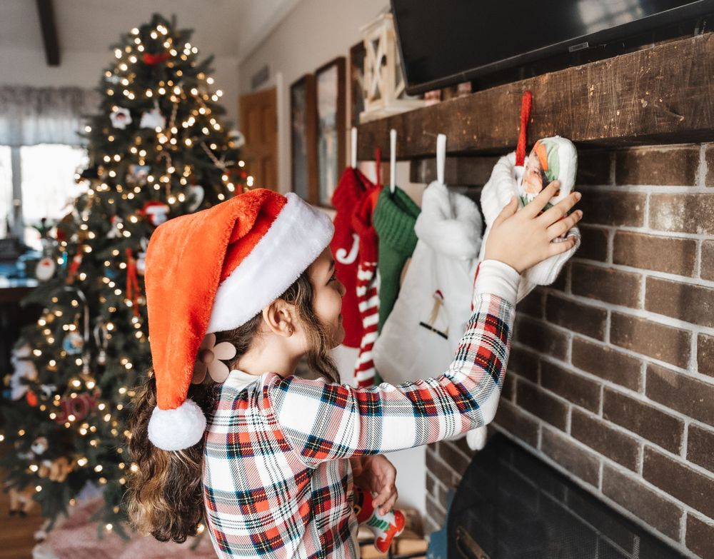 Child in plaid pajamas hangs Christmas stockings on brick fireplace with decorated tree in background.