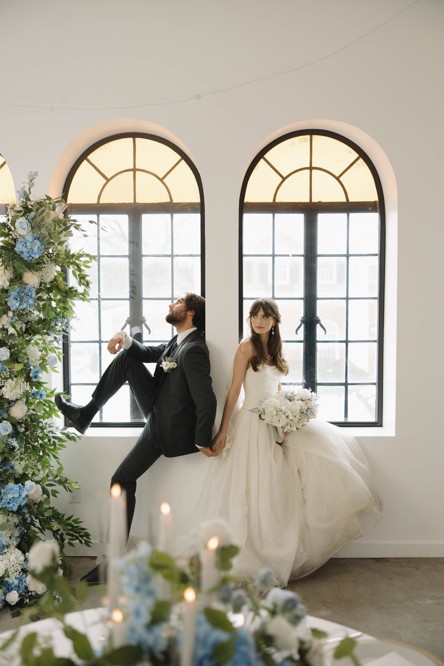 A bride and groom pose in a sunlit room with arched windows, surrounded by blue and white floral arrangements.