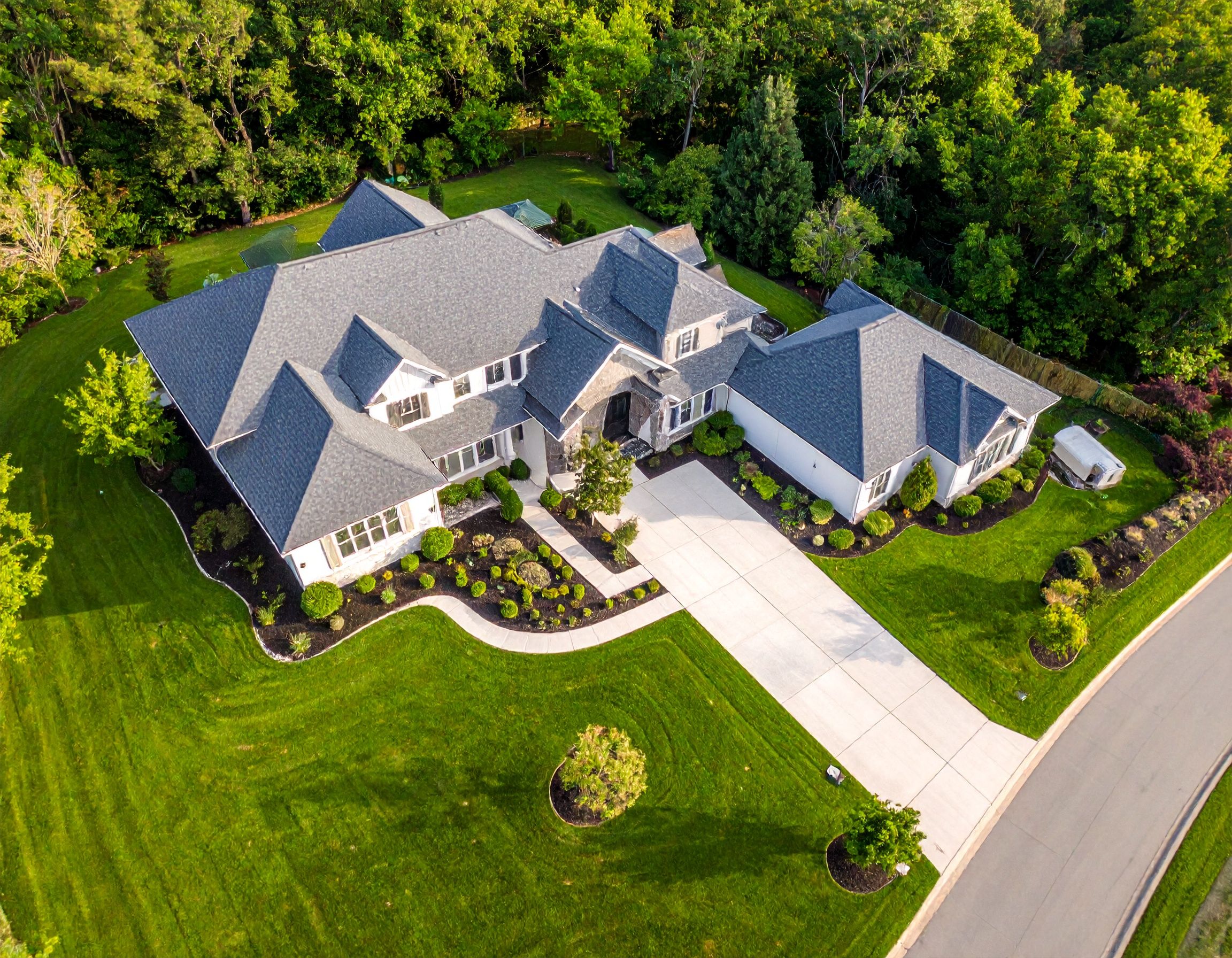 Aerial view of a white residential house with lush green landscaping and a curved driveway in a suburban neighborhood.