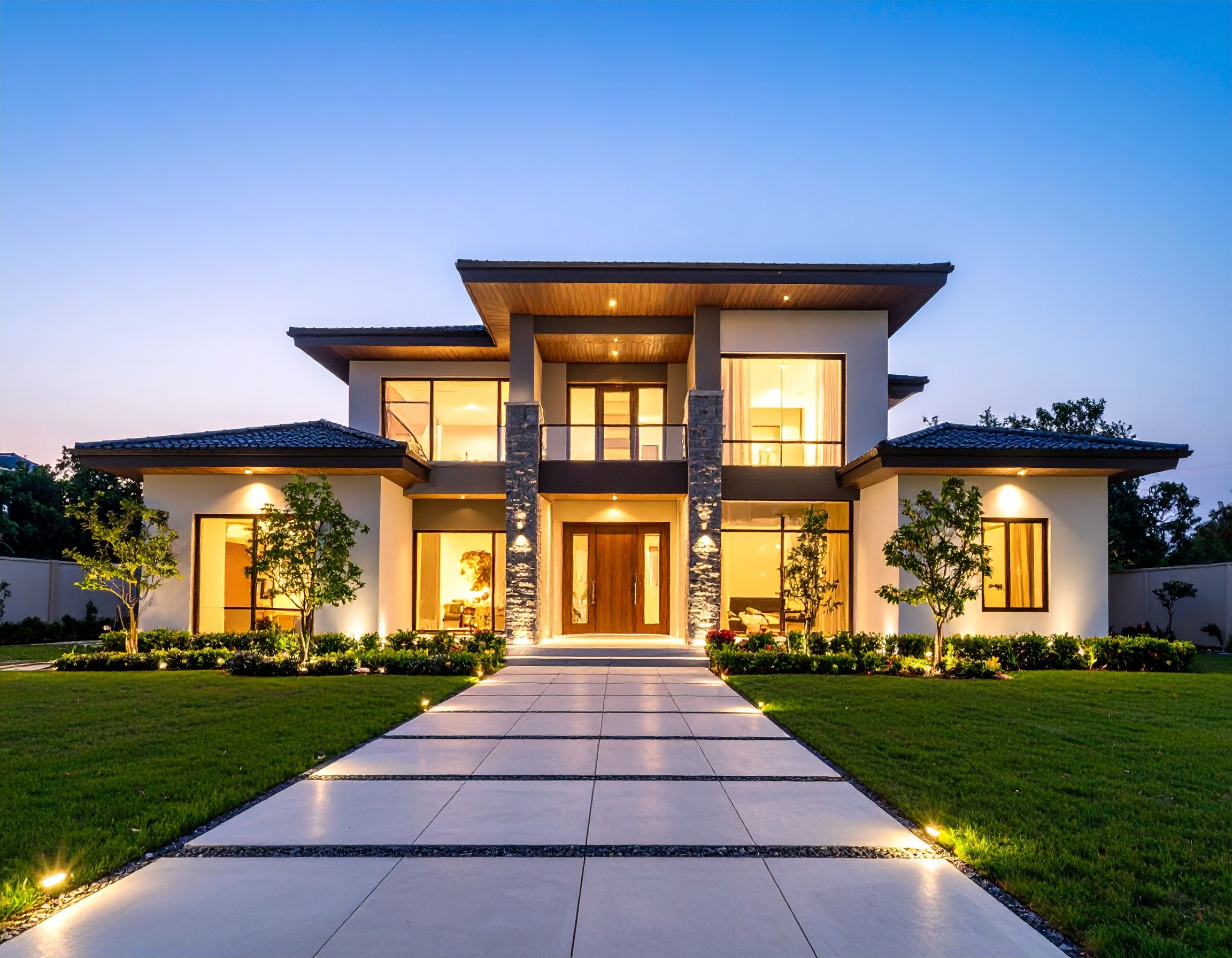 Modern luxury home exterior with illuminated driveway and large windows glowing at dusk against blue sky.
