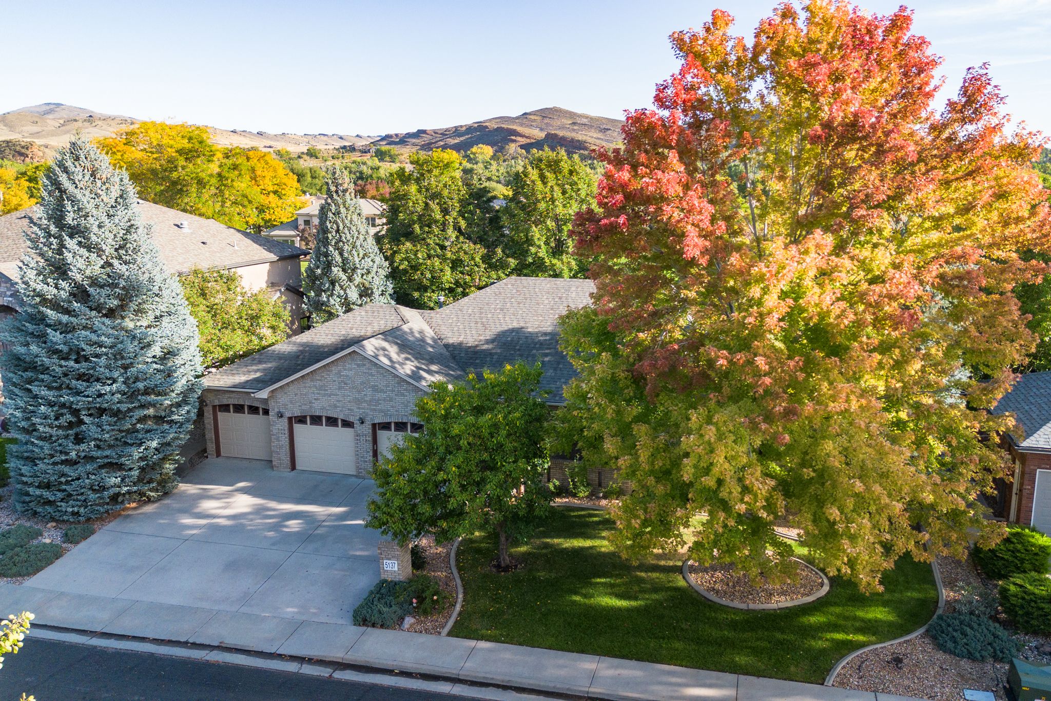 Aerial view of a suburban home with fall foliage and blue spruce trees against mountain backdrop.