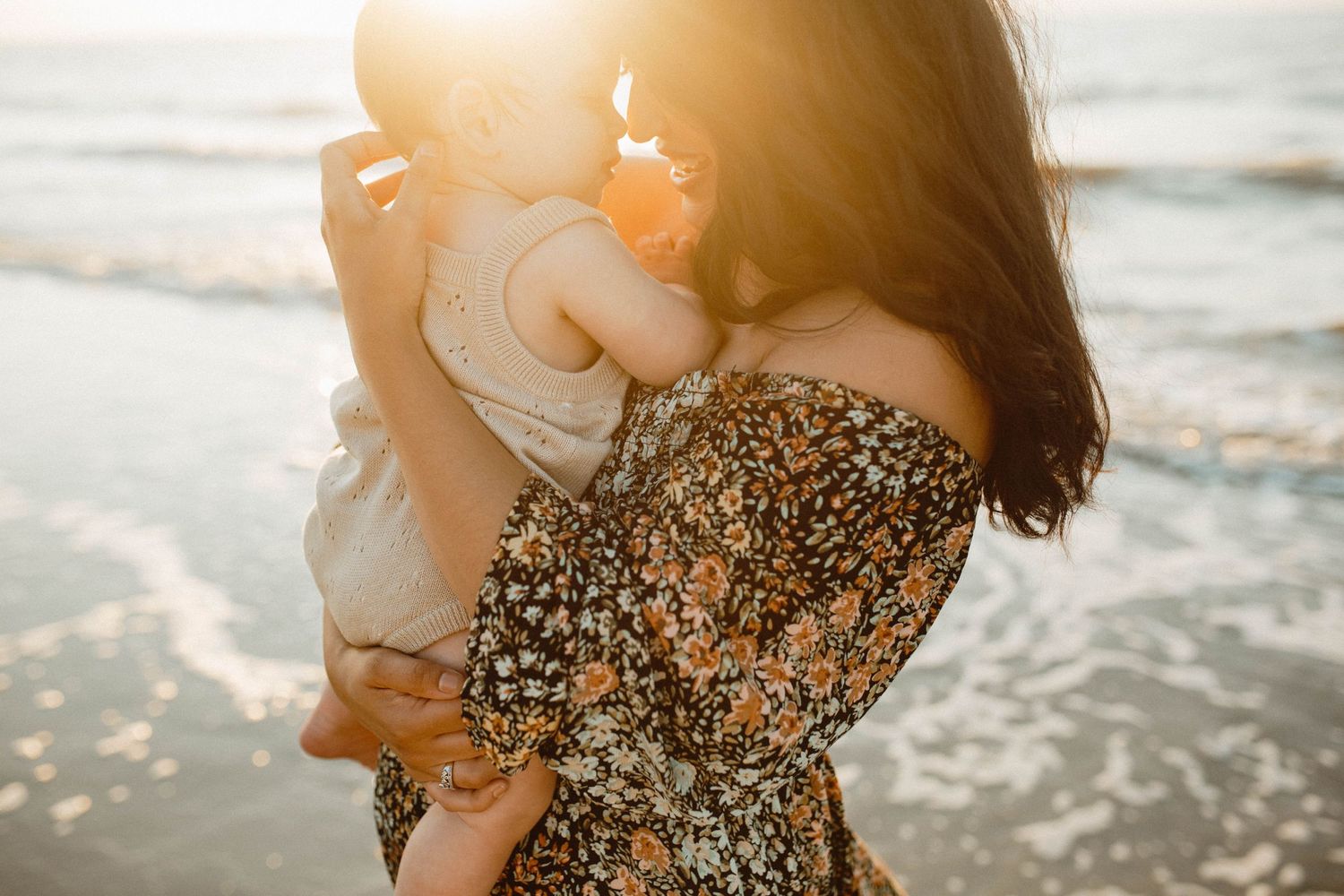 Photo by B.Logsdon of a woman in a floral dress holding a baby against a golden sunlit beach background during sunset in the Lowcountry.