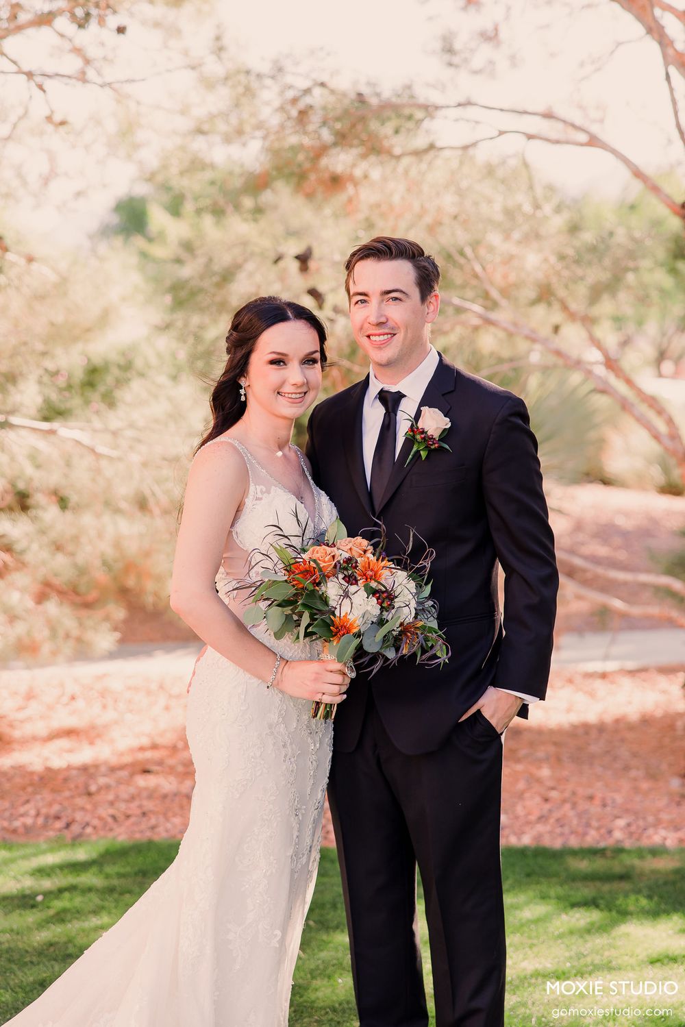 A wedding couple poses together in a desert landscape with Joshua trees and mountains in the background.