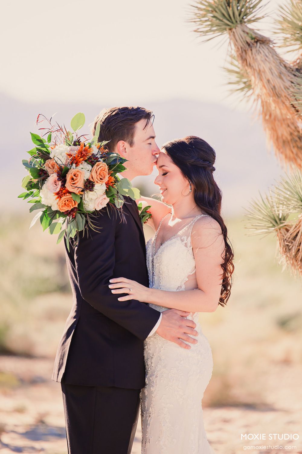 Romantic desert wedding couple embracing at sunset with coral floral bouquet and Joshua trees in background.