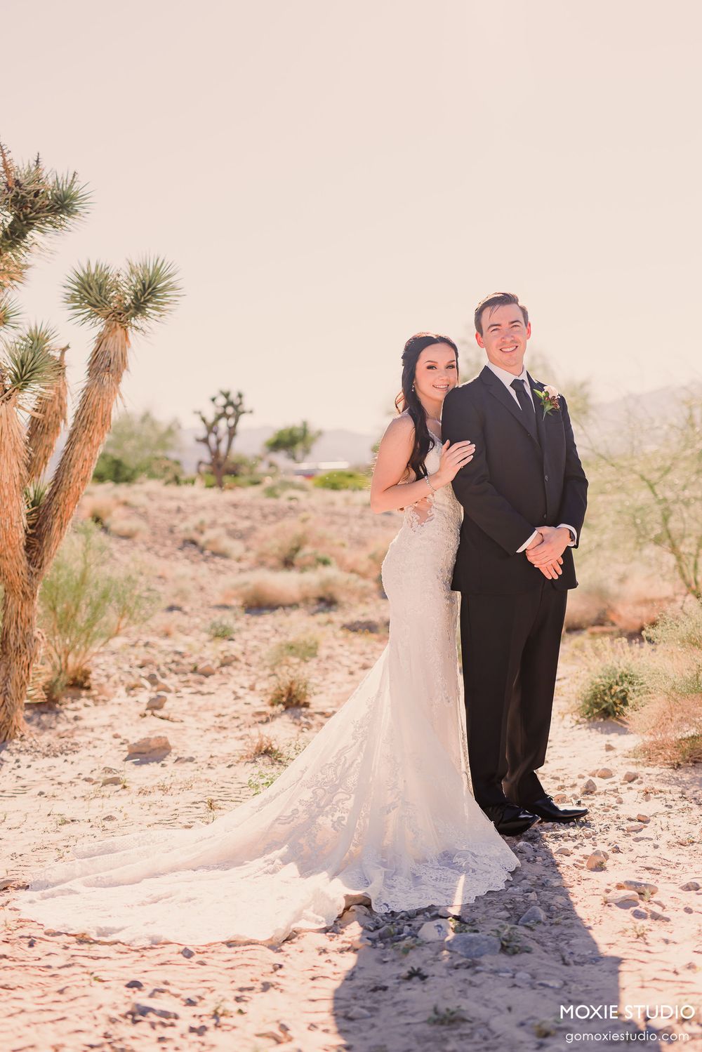 Newlyweds share tender moments in desert landscape with flowing white dress and classic black tuxedo.