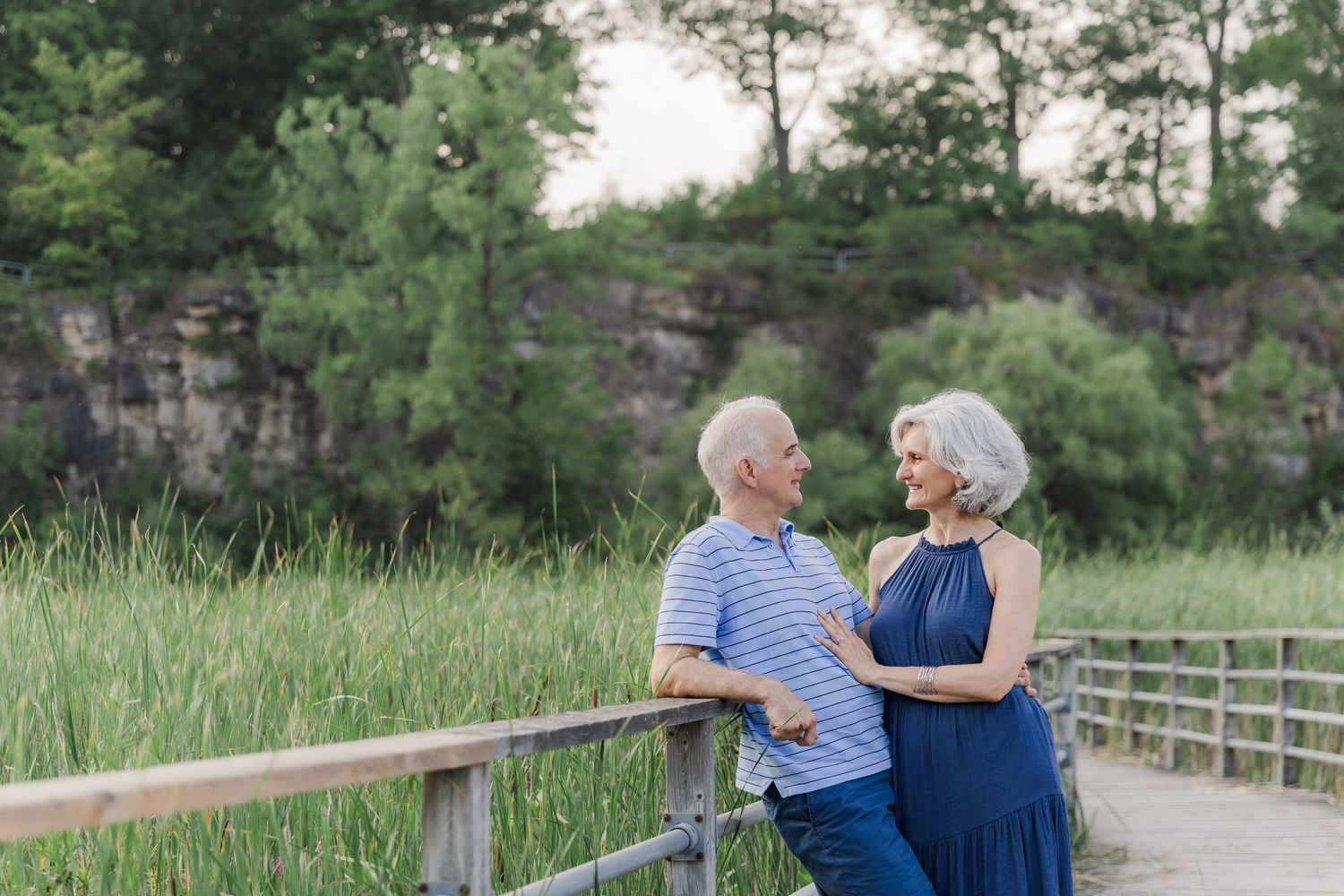Elderly couple sharing a romantic moment on wooden boardwalk near rocky cliffs and green trees at Kerncliffe Park in Burlington, Ontario.