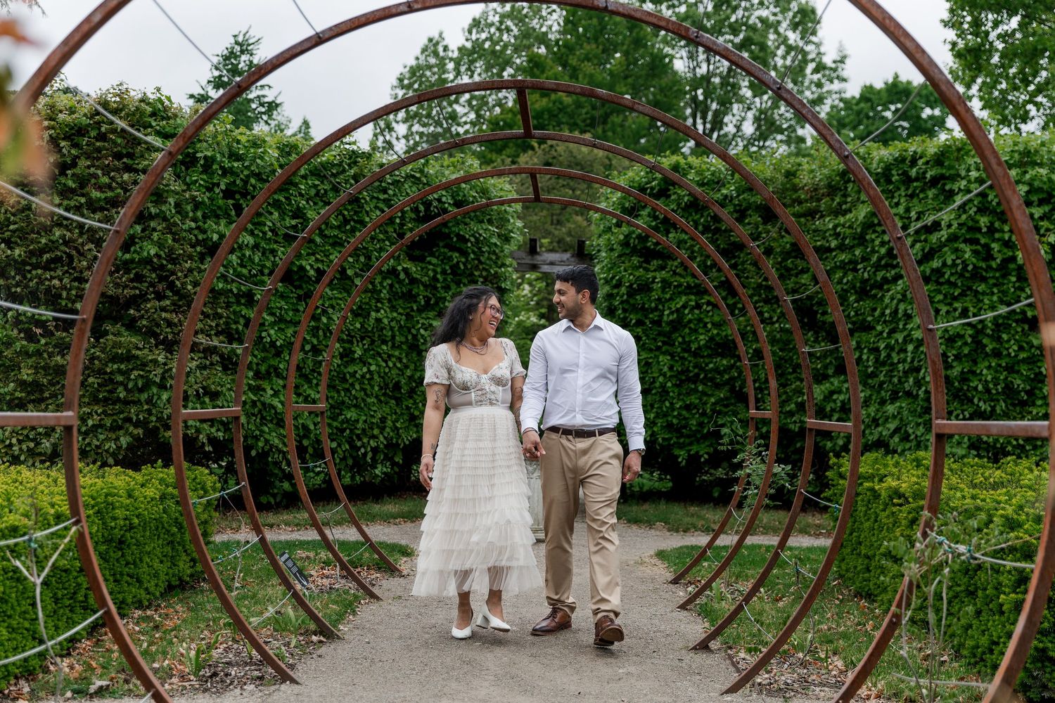 A couple walks through a curved iron garden arbor surrounded by greenery at The Guelph Arboretum.