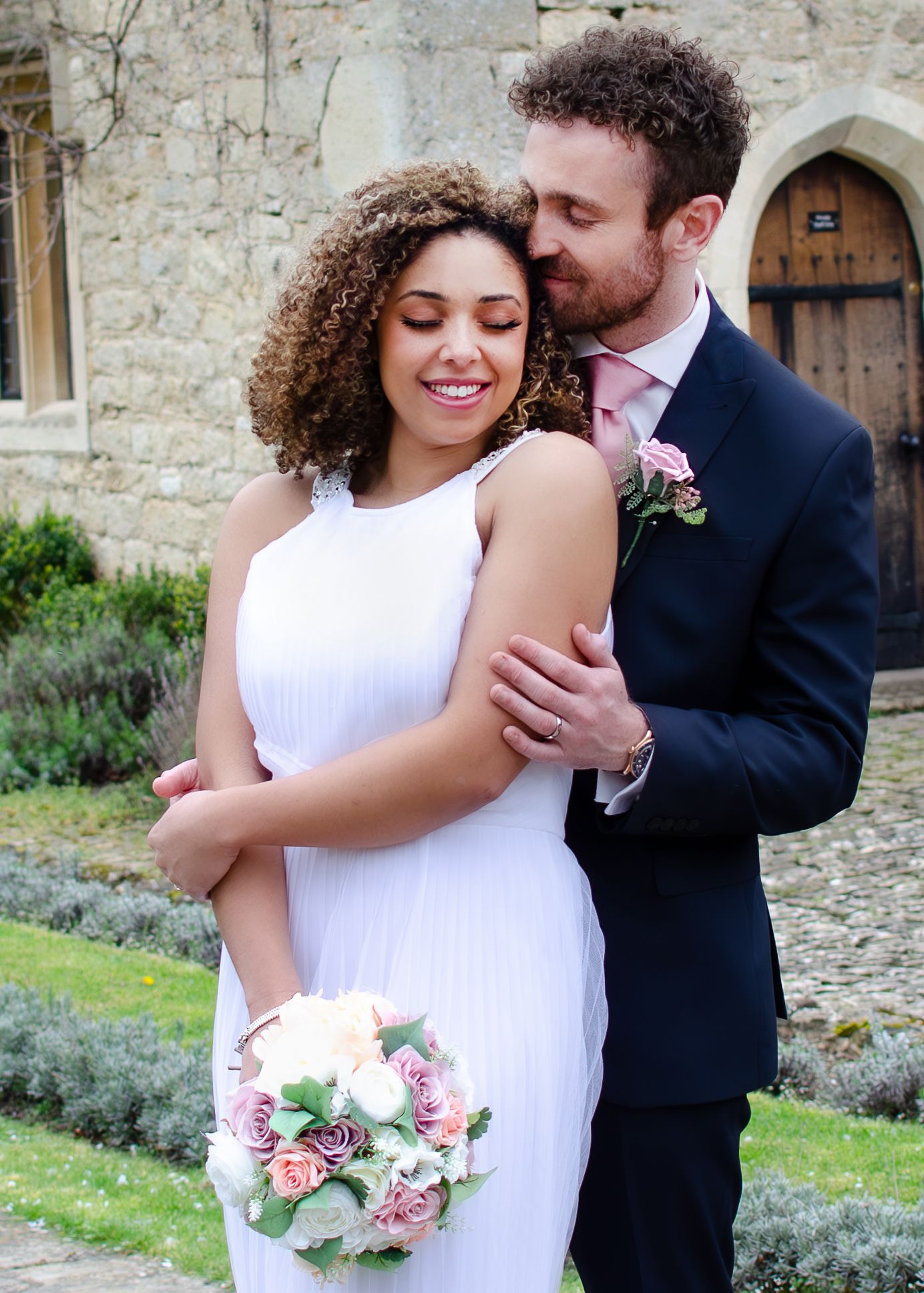 Bride and groom embracing outside a historic stone church, bride holding a pink rose bouquet.