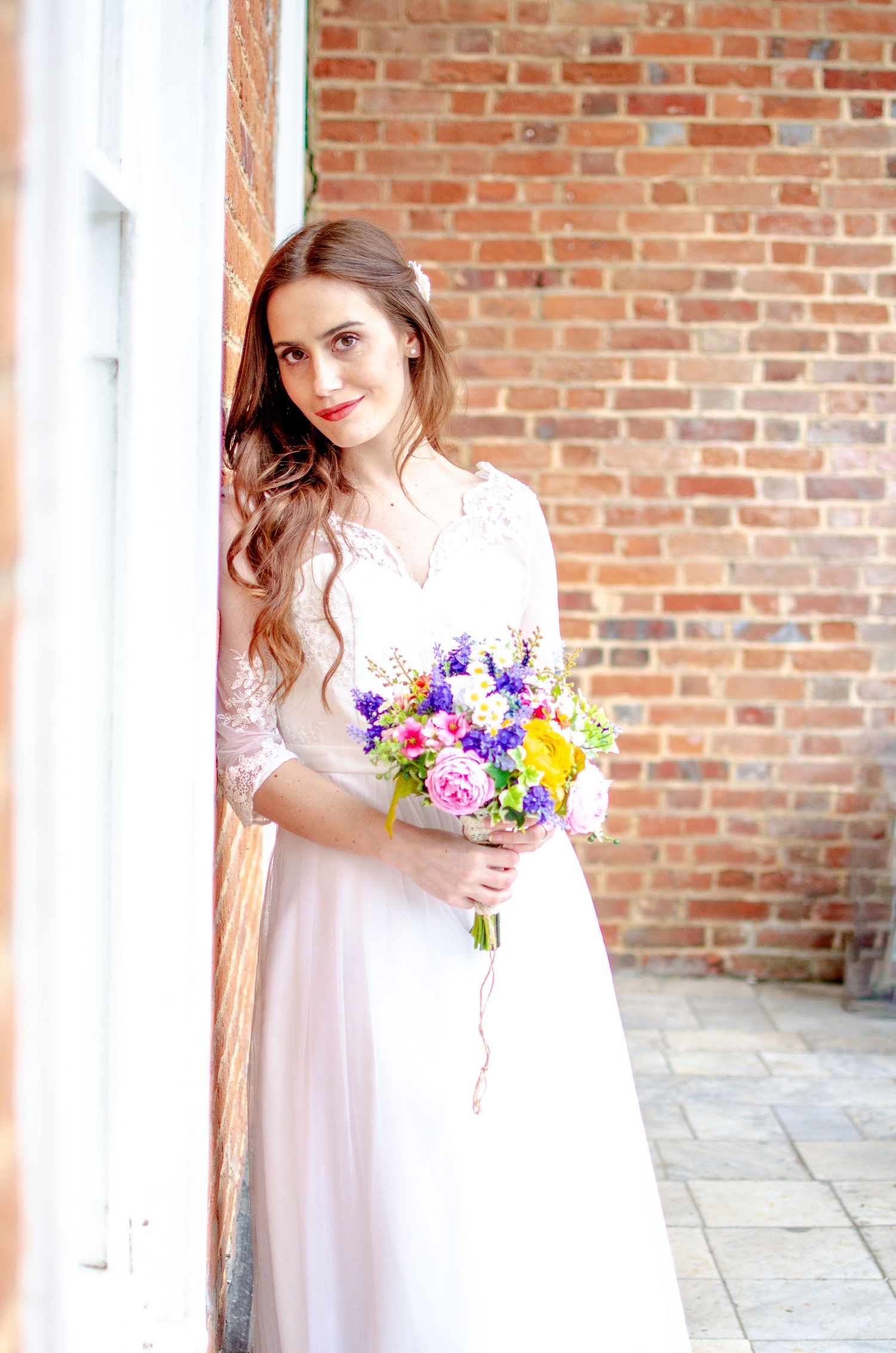 Smiling bride in lace wedding dress holding vibrant mixed flower bouquet against rustic brick wall.