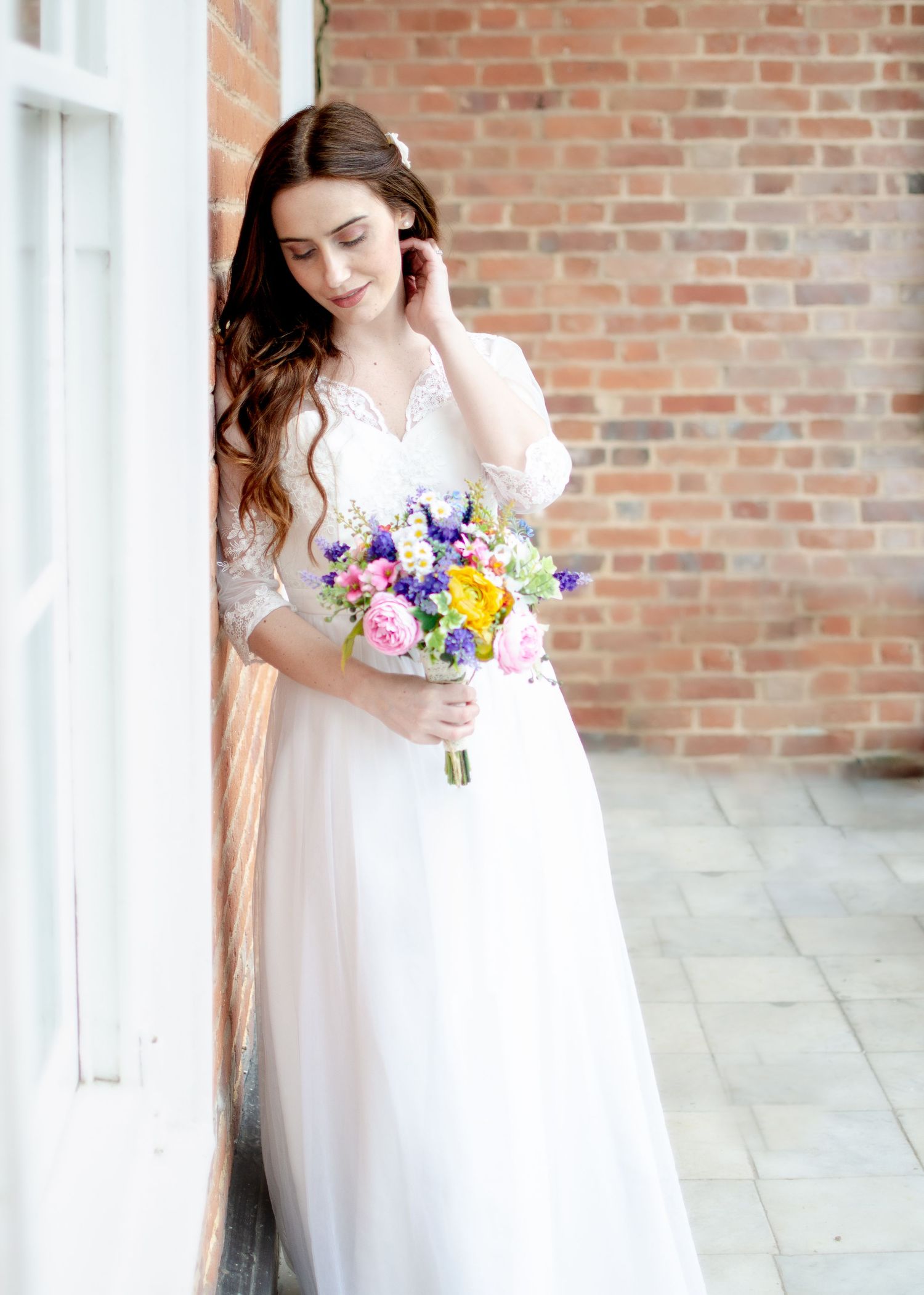 Bride in lace wedding gown holding colorful wildflower bouquet, leaning against brick wall, eyes downcast.