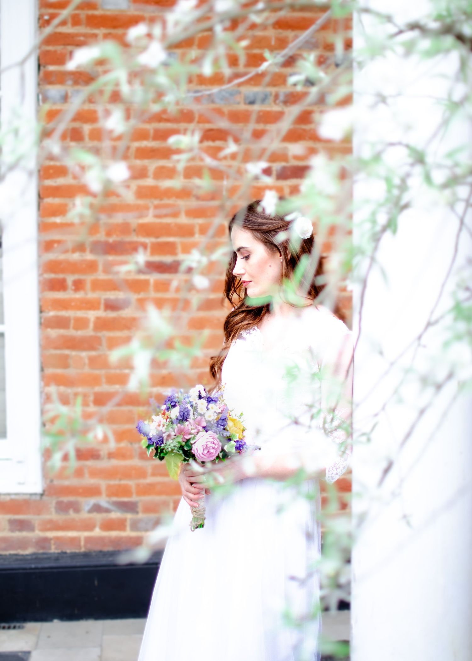 Bride in white gown holding colorful bouquet standing before red brick wall with spring branches.