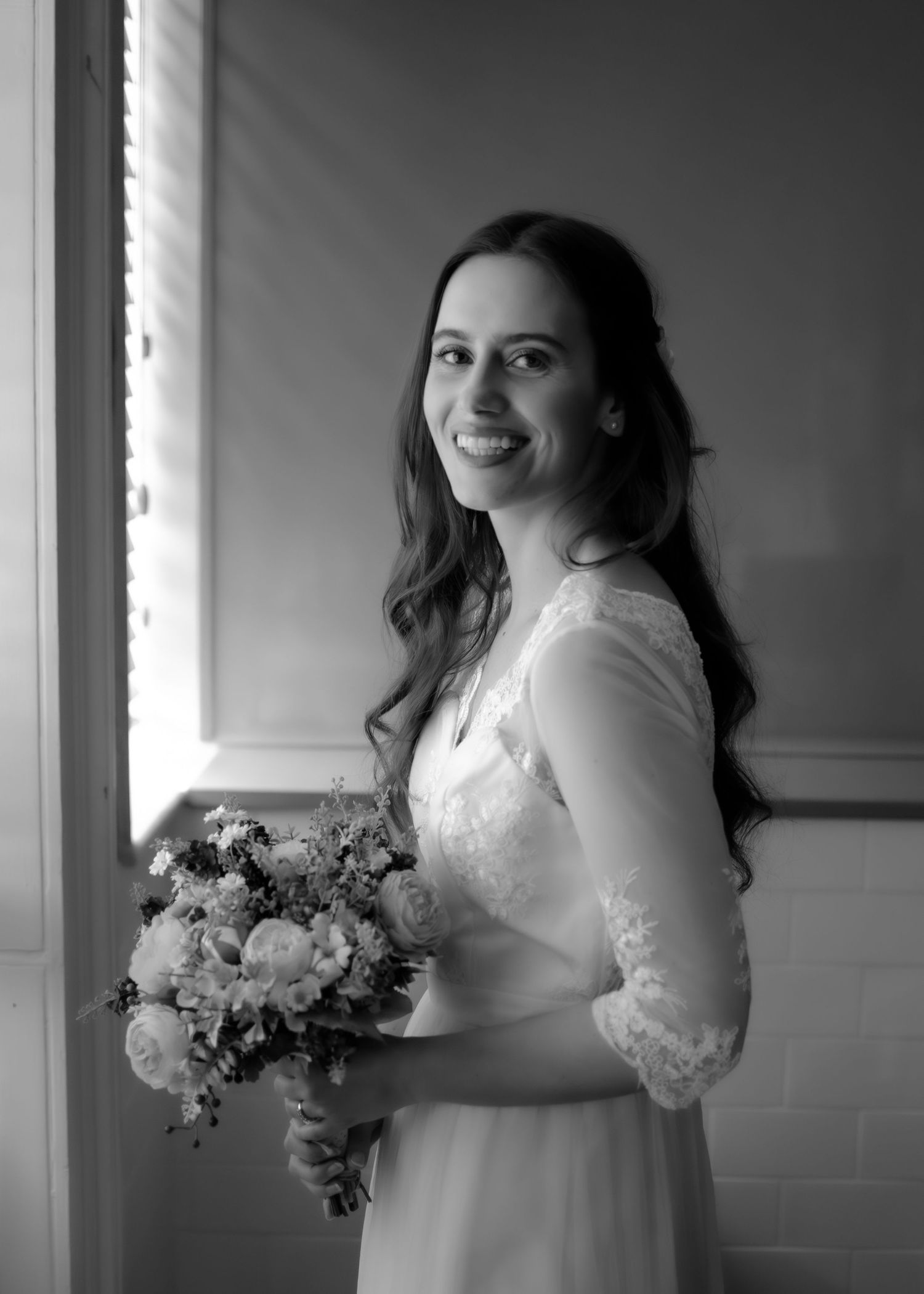 Smiling bride in lace wedding dress holding bouquet poses by window in black and white photo.
