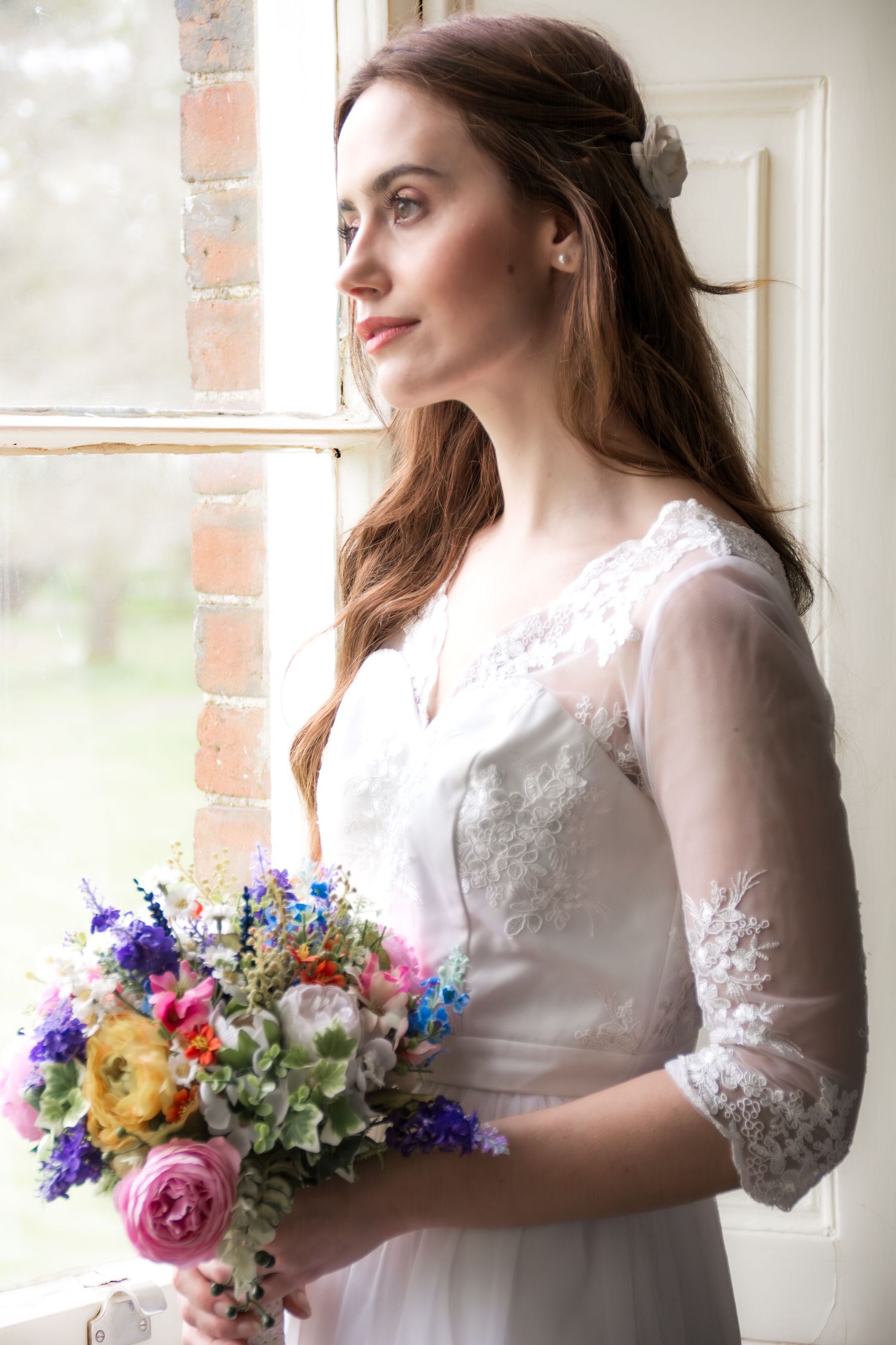 Bride in lace wedding dress gazes out a window holding a vibrant wildflower bouquet.