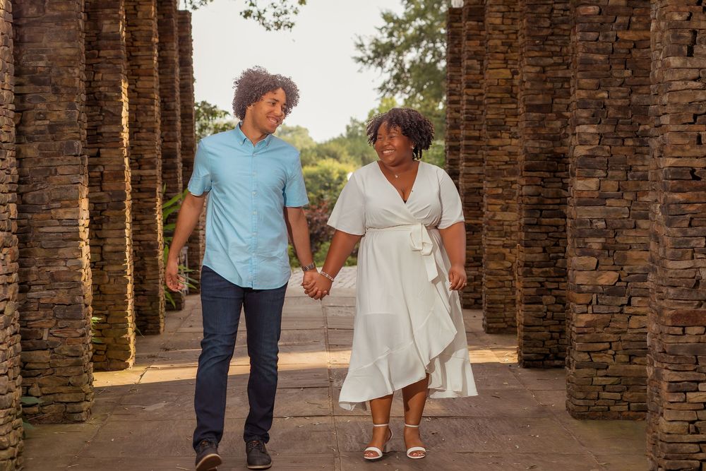 A couple holds hands while walking through a brick archway in a romantic outdoor setting.