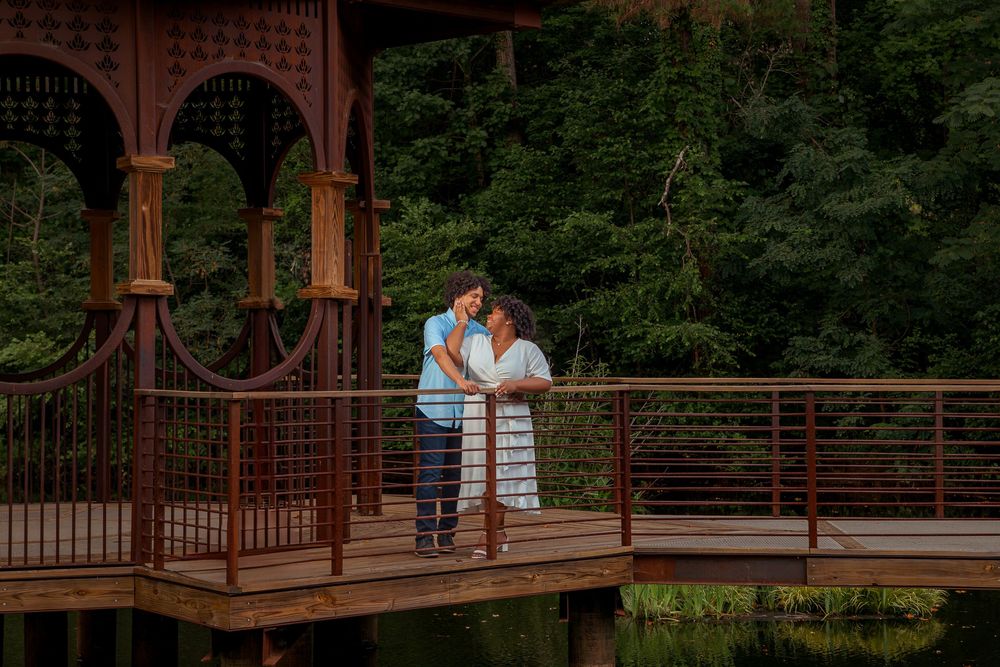 A couple shares a romantic moment on a rustic wooden bridge surrounded by lush green forest scenery.