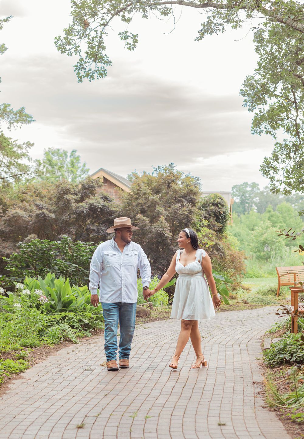 A couple walks along a curved brick pathway in a lush garden setting with trees and shrubs in the background.