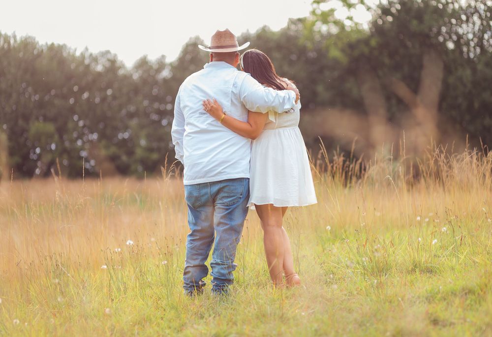 A couple embraces while standing in a golden sunlit meadow with tall grass and trees in the background.