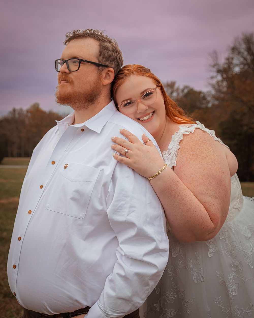 A couple in wedding attire embrace outdoors, with the person in a white dress showing off their engagement ring.