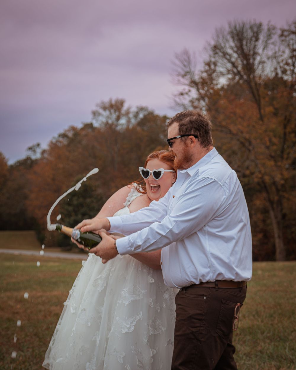 A couple wearing sunglasses sprays champagne together in an autumn field while celebrating their special day.