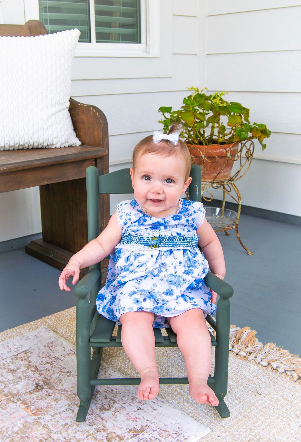 Smiling baby girl in blue floral dress sitting in a green rocking chair on a porch.