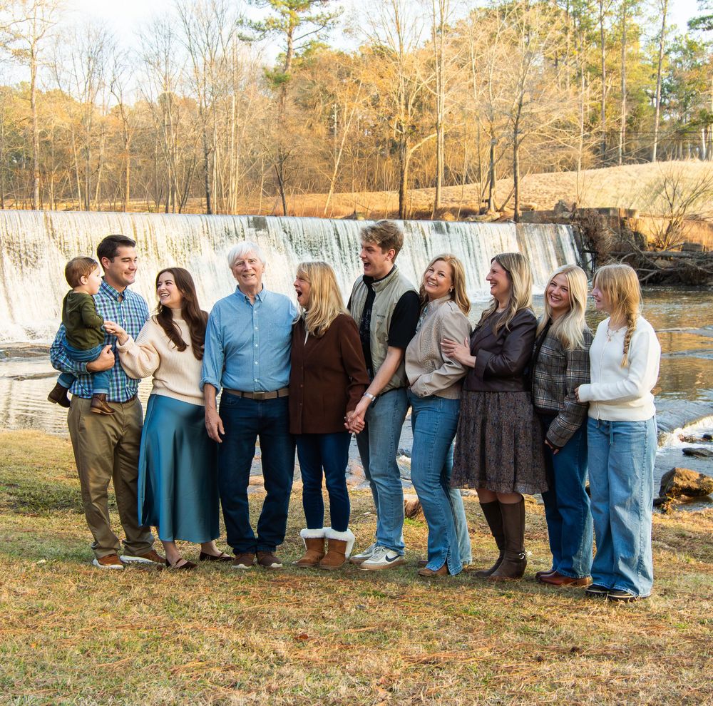 Extended family group portrait outdoors near a a waterfall in casual coordinated outfits.