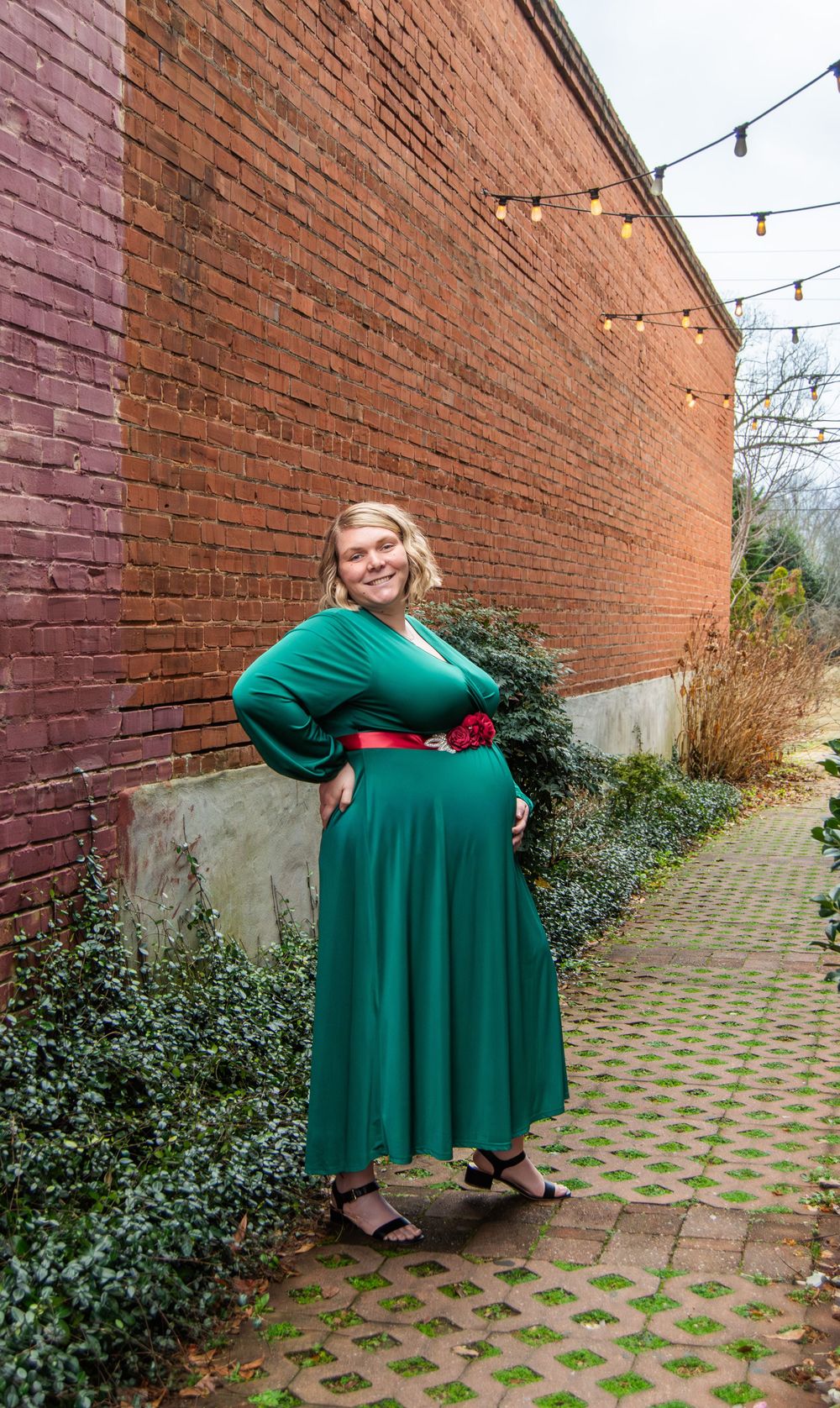 Woman in green maxi dress with red belt poses against brick wall with string lights outdoors.