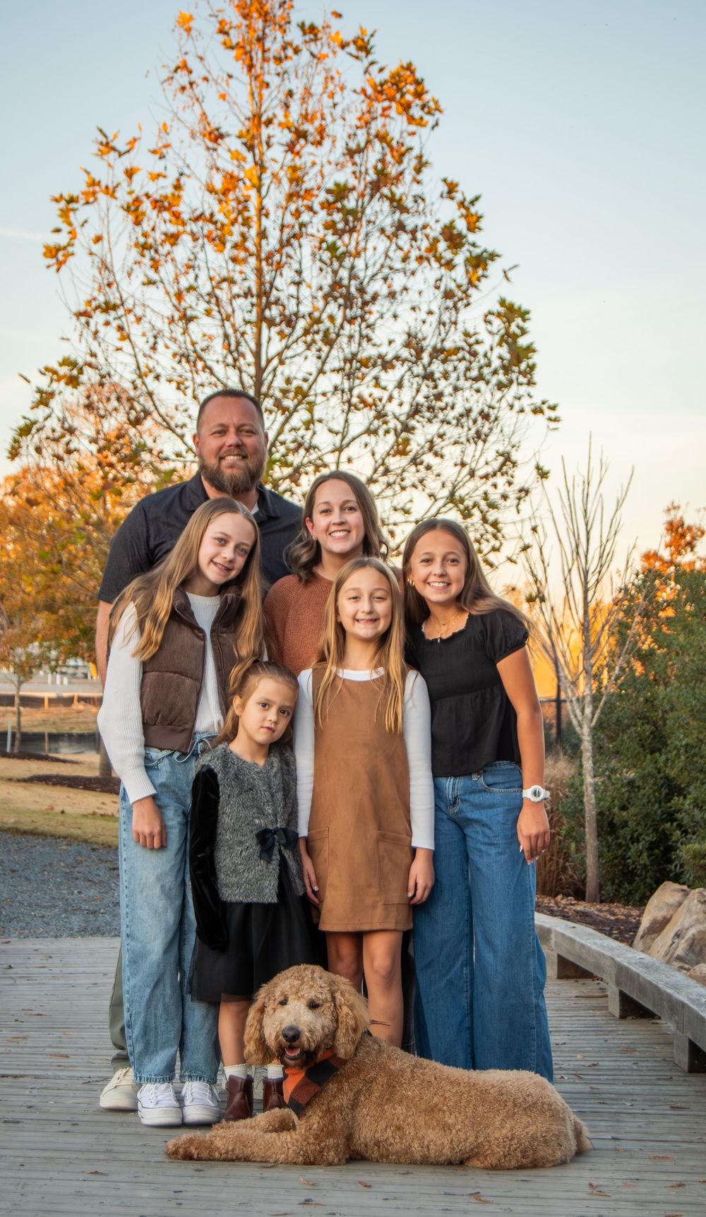 A family poses together on a wooden deck during autumn with their goldendoodle dog at sunset.