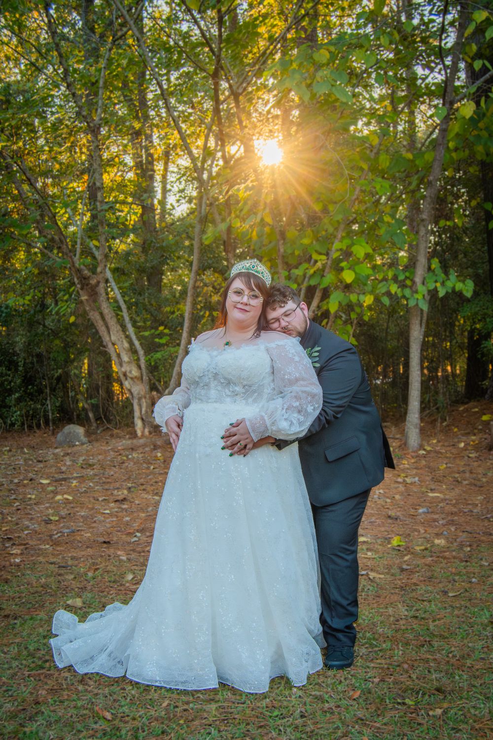 A couple in wedding attire embraces in a sunlit wooded area during golden hour as autumn leaves fall around them.