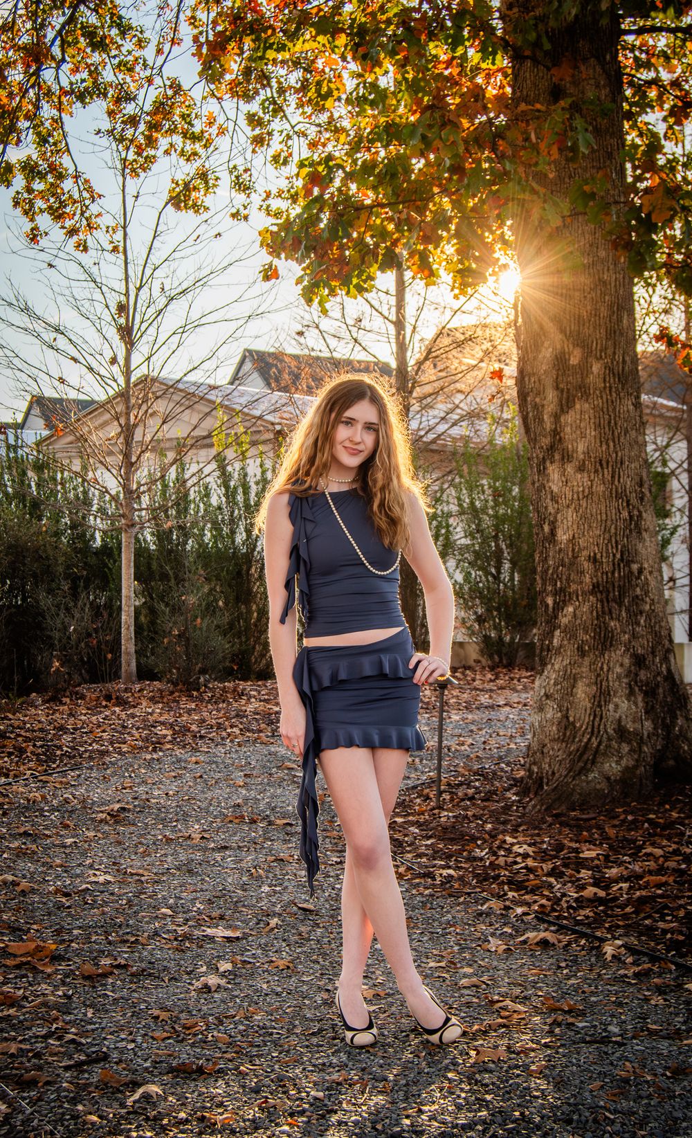 A figure in a navy dress poses in a sunlit autumn garden with fallen leaves and trees creating a warm seasonal atmosphere.