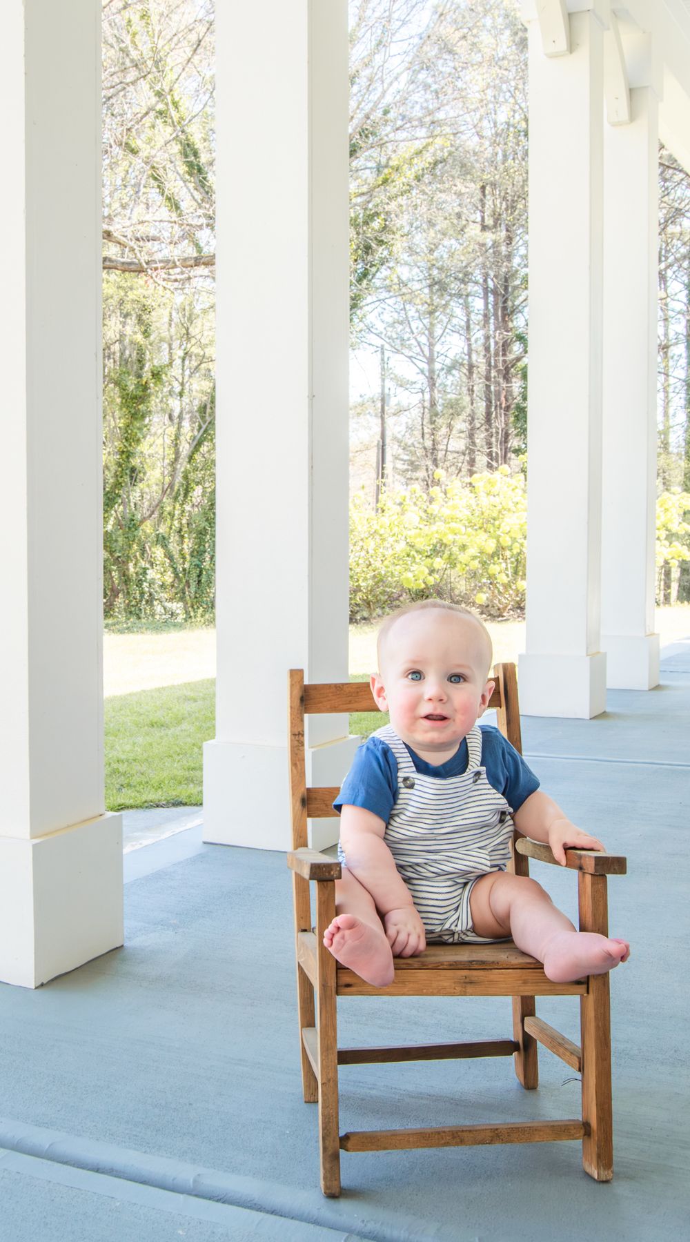 A smiling baby in striped overalls sits in a wooden chair on a white-columned porch with lush green trees behind.