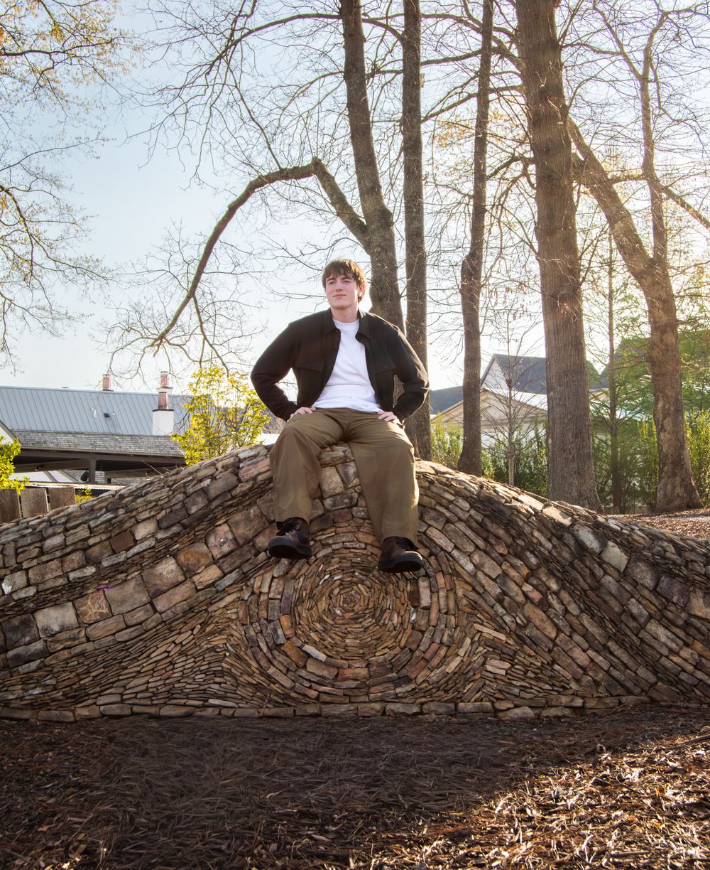 Young man sitting atop a decorative stone arch sculpture outdoors, surrounded by bare trees in a park setting.